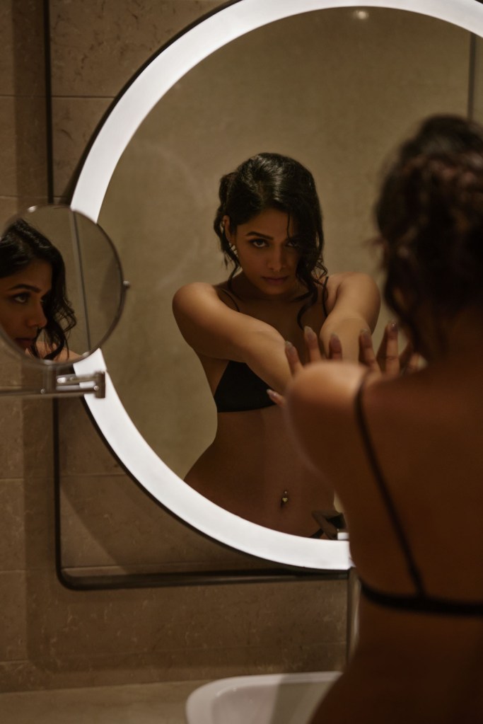 A woman in a black bikini top is looking into a circular mirror while posing in a bathroom setting.
